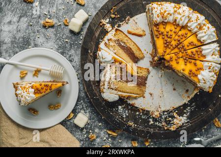 Torta su legno con pezzi tagliati sul piatto e decorazione con cioccolato bianco, noci, cocco grattugiato e panno di sacco. Su uno sfondo grigio di cemento. Foto Stock