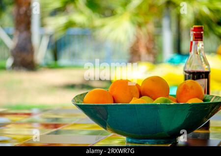 Una ciotola piena di frutta d'arancia e una bottiglia di vino su un tavolo colorato in un ambiente esotico. Splendido resort estivo con palme sullo sfondo. Foto Stock