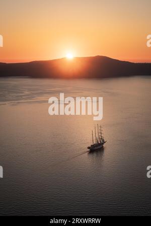 La baia al largo di Thera, Santorini, con il sole che tramonta dietro l'isola di Thirasia, il cielo limpido e dorato e la barca a vela alberata nel mezzo della baia Foto Stock