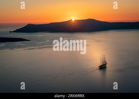 La baia al largo di Thera, Santorini, con il sole che tramonta dietro l'isola di Thirasia, il cielo limpido e dorato e la barca a vela alberata nel mezzo della baia Foto Stock