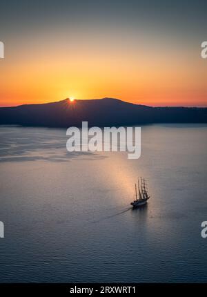 La baia al largo di Thera, Santorini, con il sole che tramonta dietro l'isola di Thirasia, il cielo limpido e dorato e la barca a vela alberata nel mezzo della baia Foto Stock