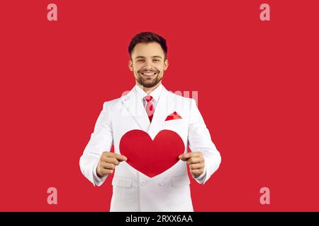 Elegante uomo barbuto in abito classico bianco con cravatta rossa che regge il cuore rosso per san valentino e sorride isolato su sfondo rosso in studio Foto Stock