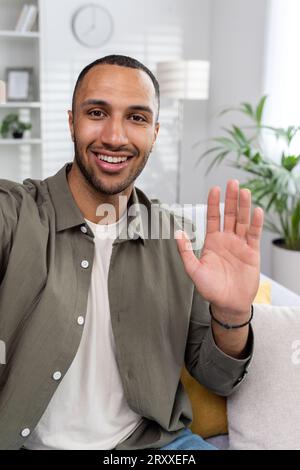 Foto verticale. Un giovane musulmano sorridente sta trasmettendo un video online da casa. Sedersi sul divano e salutare la telecamera. Foto Stock