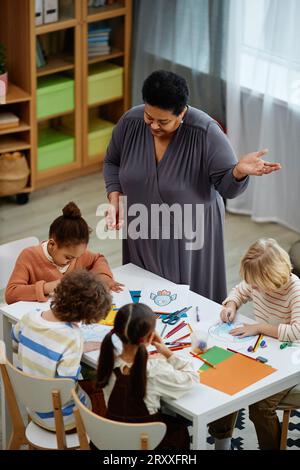 Ritratto verticale di donna matura nera come insegnante femminile che aiuta i bambini piccoli a lezione d'arte e di artigianato in età prescolare Foto Stock