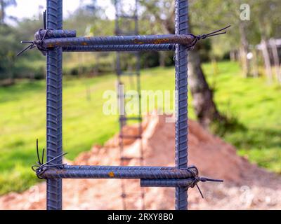 Fotografia ravvicinata delle barre di ferro strutturali e delle fascette per le colonne di cemento di una costruzione rurale, con una pila di sabbia sul retro. Foto Stock
