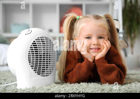 Una bambina carina che si scalda vicino al riscaldamento elettrico della ventola in camera da letto Foto Stock