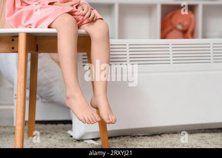 Una bambina scalza che scalda le gambe vicino al radiatore a casa Foto Stock