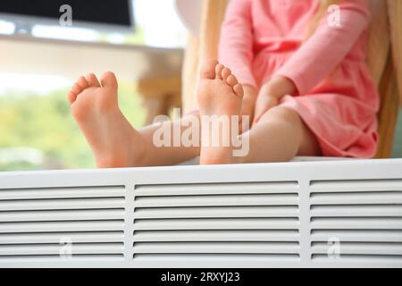 Bambina a piedi nudi che scalda le gambe vicino al radiatore a casa, primo piano Foto Stock