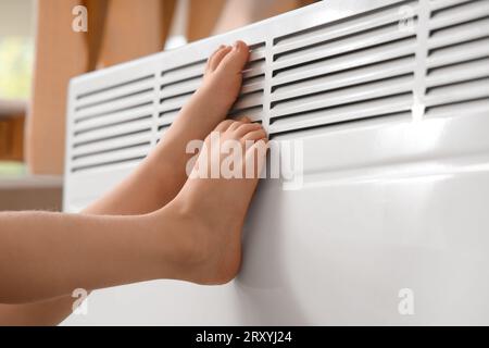 Bambina a piedi nudi che scalda le gambe vicino al radiatore a casa, primo piano Foto Stock