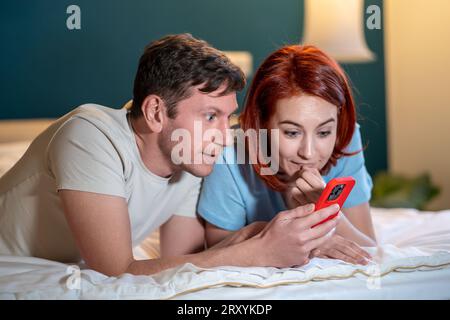 Felicissima coppia europea sdraiata sul letto insieme Guarda lo schermo dello smartphone sorpreso vincere la lotteria online Foto Stock