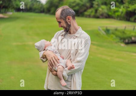 Un felice padre di 40 anni culla il suo neonato in un parco soleggiato. Amore, famiglia e generazioni in armonia Foto Stock