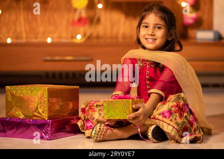 Bambina indiana allegra che tiene in mano delle confezioni regalo per celebrare Diwali mentre è seduta sul pavimento, Raksha Bandhan, concetto Bhai Dooj. Foto Stock