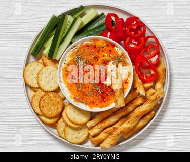 Salsa di formaggio gelatina al peperoncino caldo in ciotola servita con cracker, grissini salati, pepe fresco e cetriolo su piatto su un tavolo di legno bianco, vista orizzontale Foto Stock