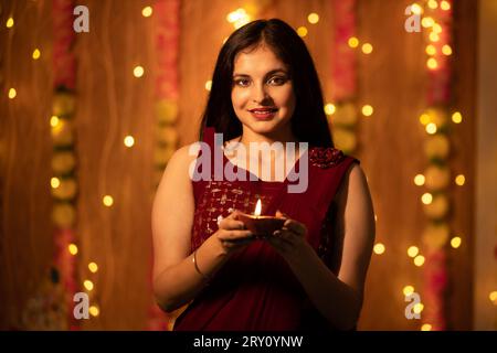 Ritratto di una bella giovane donna indiana sorridente in abito tradizionale che tiene una lampada ad olio o un diya con decorazioni di luci festive nello sfondo Foto Stock
