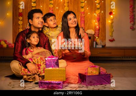 Felice giovane famiglia indiana in abito tradizionale con un sacco di regali seduti sul pavimento per celebrare il festival diwali a casa. Foto Stock