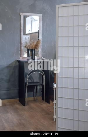 Camera moderna arredata con schermo pieghevole bianco, specchio rettangolare appeso su un tavolo nero dove poggia un vaso di pampas beige. Comoda PL Foto Stock