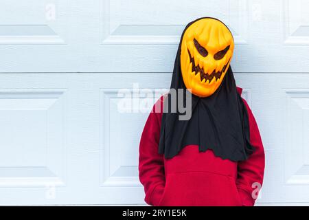 Una bambina che indossa una spaventosa maschera di zucca di Halloween, ha lasciato uno spazio a sinistra, appoggiandosi contro la porta bianca di un garage. Concetto di celebrazione, perche' Foto Stock