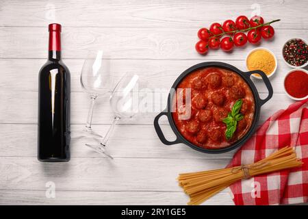 Polpette in salsa di pomodoro in una padella con ciliegia, pomodori, bottiglia di vino e due bicchieri su una tavola di legno bianca. Vista dall'alto. Foto Stock