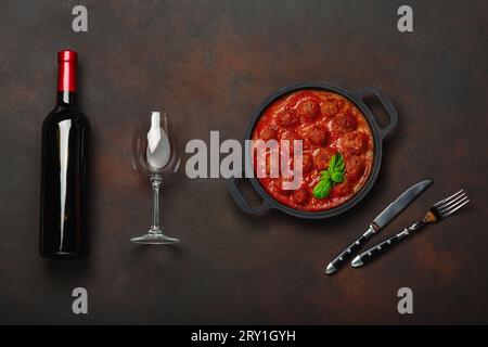Polpette in salsa di pomodoro con spezie, pomodori ciliegini, pasta e basilico in padella con bottiglia di vino e clessidra su fondo marrone arrugginito. T Foto Stock