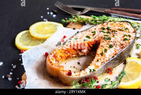 Salmone al forno con erbe aromatiche e limone su carta da forno, vista dall'alto. Bistecca di salmone fritta con timo e limone servita su un piatto di legno su un tavolo nero. Foto Stock
