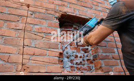 Un costruttore con una T-shirt nera e guanti che scalpellano la muratura di una casa con un trapano pneumatico, lavorando per distruggere un muro di mattoni Foto Stock