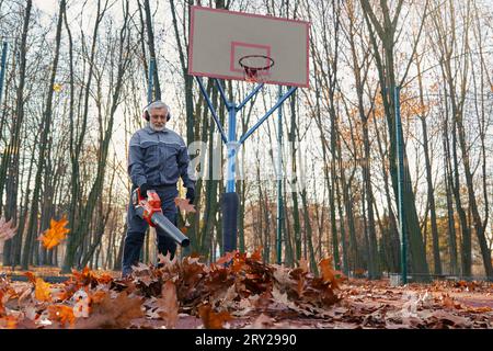 Addetto alla manutenzione barbuto che utilizza un soffiatore per pulire il campo da basket abbandonato. Vista angolare ridotta dell'uomo anziano in uniforme per rimuovere foglie asciutte con soffiatore all'aperto. Concetto di lavoro stagionale. Foto Stock