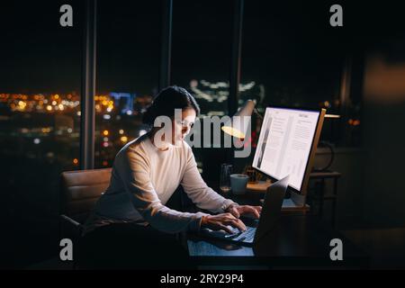 Donna d'affari dedicata lavora fino a tardi, digitando il suo laptop alla scrivania dell'ufficio. Imprenditrice donna che lavora fino a tardi, concentrata e determinata all'incontro Foto Stock