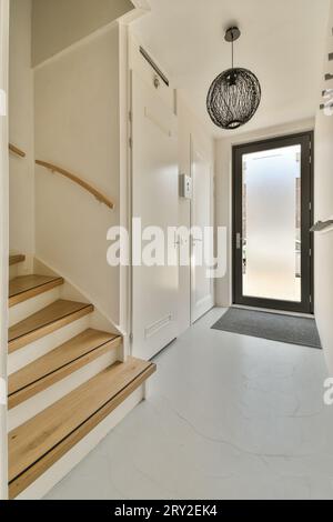 Corridoio stretto che porta verso la porta con scale e lampada appesi al soffitto in un moderno appartamento con parete e pavimento bianchi Foto Stock