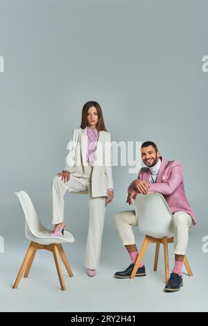 uomo gioioso con blazer lilla seduto e guardando la fotocamera vicino a una donna che posa con la poltrona grigia Foto Stock