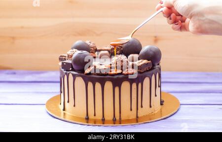 La mano di una donna versa una salsa al caramello salata sulla torta al cioccolato, decorata con dolci, tartufi, biscotti e noci Foto Stock