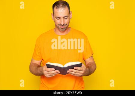 Bearded Hispanic man nei suoi 40 anni, che si allenava con la mente e si divertiva a leggere un libro, isolato sullo sfondo giallo dello studio. Foto Stock