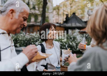 Donna anziana sorridente che si gusta spuntini con gli amici mentre si trova in strada Foto Stock