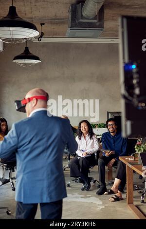 Colleghi sorridenti che guardano un uomo d'affari maturo che indossa la realtà virtuale in ufficio Foto Stock