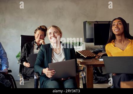 Felici colleghi d'affari seduti con i computer portatili mentre si trovano seduti in un ufficio creativo durante una riunione Foto Stock