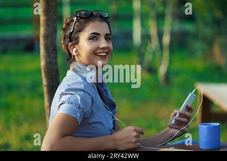 All'aperto, giovane donna lavora dinamicamente, gestendo telefono e laptop. In camicetta a righe, i suoi capelli sono tenuti da occhiali, in mezzo al paesaggio montano. Il suo expressi Foto Stock