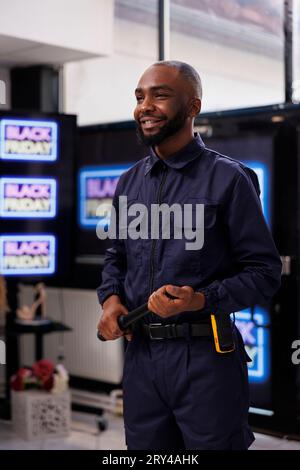 Una guardia di sicurezza afroamericana sorridente che indossa un'uniforme che tiene il bastone in mano all'ingresso del centro commerciale contro gli striscioni al neon luminosi. Apertura del negozio al dettaglio il Black Friday Foto Stock