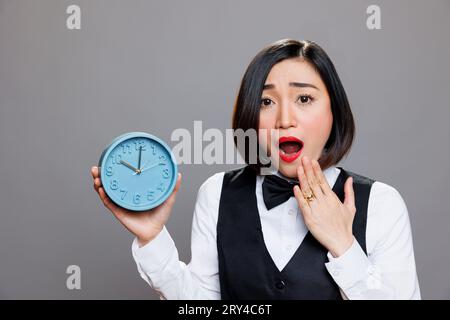 Scioccata cameriera asiatica che teneva la sveglia e guardava la macchina fotografica mentre copriva la bocca aperta. Giovane donna ansiosa receptionist che mostra tempo mentre esegue un ritratto in ritardo Foto Stock