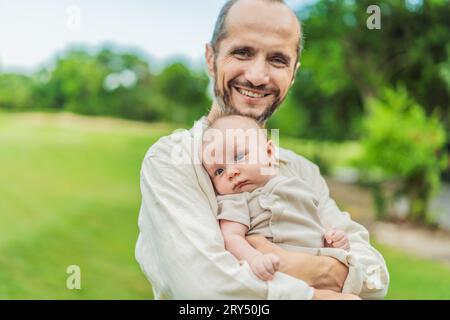 Un felice padre di 40 anni culla il suo neonato in un parco soleggiato. Amore, famiglia e generazioni in armonia Foto Stock