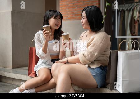 Una giovane donna asiatica affascinante e allegra ama parlare chiacchierare e sorseggiare un caffè mentre riposa sulle scale con la sua amica durante una sh Foto Stock