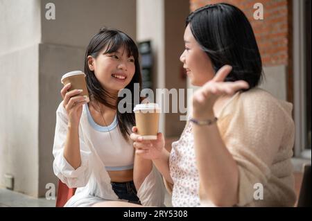 Una bella giovane donna asiatica ama parlare e bere un caffè con la sua amica mentre si siede sulle scale della città. Stile di vita urbano Foto Stock