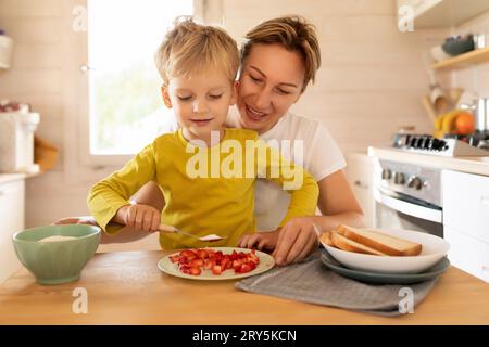 Piccolo figlio biondo caucasico e madre che preparano la colazione con fragole mentre sono seduti in cucina Foto Stock