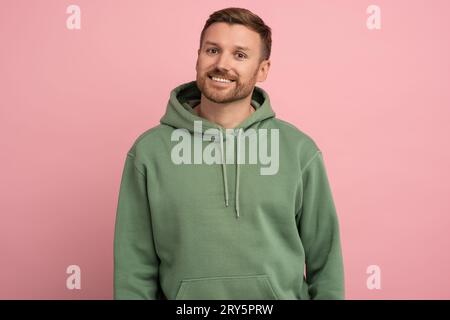 Ritratto uomo barbuto caucasico con felpa verde che guarda la fotocamera sorridente su sfondo rosa. Foto Stock