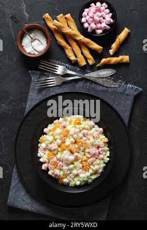 Insalata Ambrosia con ananas, arance mandarine, yogurt, mini marshmallow, cocco e panna montata in una ciotola nera su un tavolo di cemento con ingredienti Foto Stock