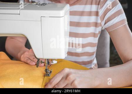 Cucitura a mano di tessuti gialli su una moderna macchina da cucire sul luogo di lavoro in atelier. Le mani delle donne cuciscono pezzi di tessuto sul primo piano della macchina da cucire. H Foto Stock