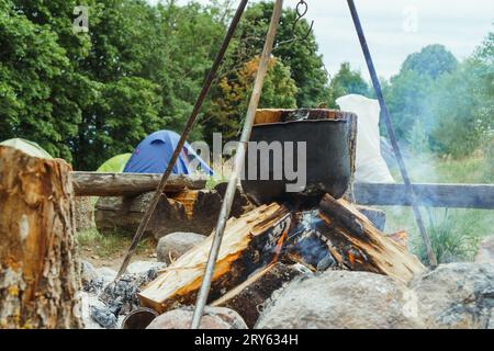 Processo di cottura del pasto in calderone metallico appeso al treppiede sopra la legna da ardere, in piedi contro le tende e la foresta verde. Preparare il cibo al falò per il campo Foto Stock