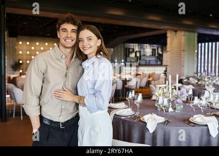 coppia felice innamorata che guarda la macchina fotografica nella sala banchetti con tavoli festosi decorati, giorno speciale Foto Stock