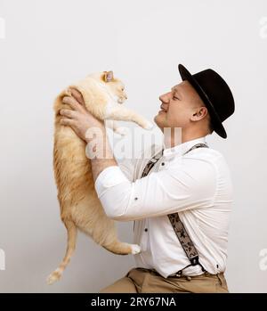 Un uomo con una camicia e un cappello regge un grosso gatto, sfondo grigio Foto Stock