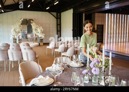 felice manager di eventi sorridente vicino al tavolo con ambiente festivo, fiori e candele nella sala eventi Foto Stock