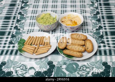 Due piccole ciotole di Hummus e guacamole fatti in casa accompagnate da piatti con pane tostato integrale e bastoncini di pane tostato su un elegante panno Tablecloth Ador Foto Stock