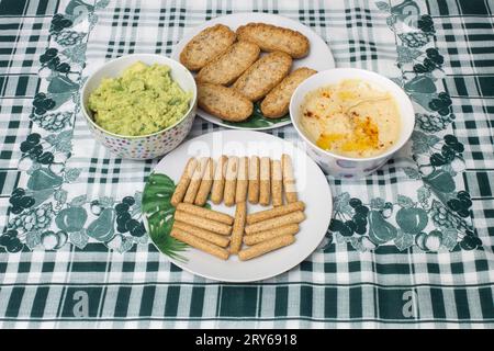 Due ciotole di hummus cremoso e salsa guacamole sono poste su un tavolo con una tovaglia a foglia e geometrica, insieme a toast di grano intero e grissini Foto Stock